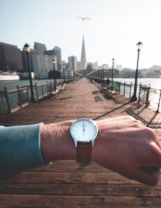 An arm with a watch on the wrist; person is stood on a boardwalk next to a river