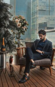 Wealthy-looking young man sat on a deck typing into a computer. Next to him is a bottle of Champagne.
