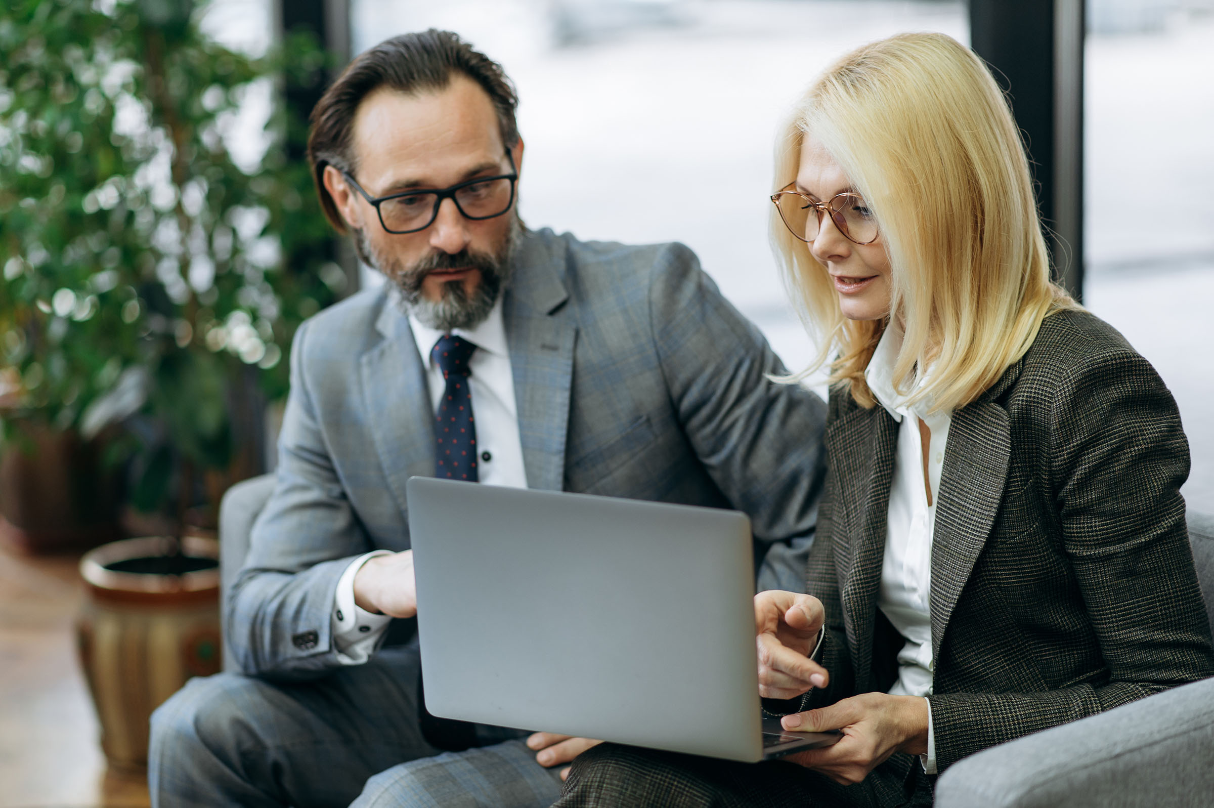 Man and woman in business setting look at a laptop screen. Woman is blonde, man has brown hair and both have glasses